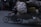 A motorcycle carries a large, overloaded pile of burlap sacks and various items tightly bound together with cords. The rider, wearing a helmet, navigates through an urban street. The scene is slightly dim, indicating possible dusk or evening lighting.