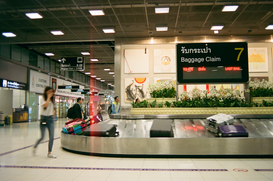 The image depicts a busy airport baggage claim area with several suitcases and bags scattered around, indicating travelers arriving or departing. A luggage carousel is prominently visible on the right side of the frame, with multiple suitcases and bags attached to it. The scene also includes people walking by in various directions, some carrying backpacks and handbags as they navigate through the airport.