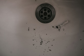 A dirty sink with scattered ashes or debris around the drain and a used cigarette filter lying nearby.