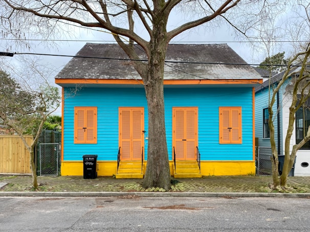 A vibrant house painted in bright turquoise blue with contrasting orange shutters and yellow steps. The house features three sets of shutters and a narrow porch. A leafless tree is prominently located in front of the house, intersecting the facade. The surrounding includes a paved sidewalk and another house painted in a more muted color scheme, visible on the right.