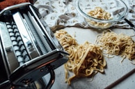 A close-up view of freshly made pasta on a countertop, featuring a pasta machine on the left and several piles of different types of pasta, including ribbons and noodles. A glass bowl with additional pasta is visible in the background, along with a decorative cloth.