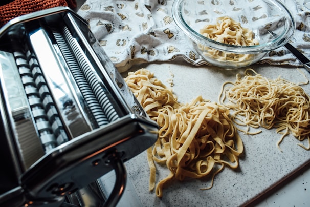 A close-up view of freshly made pasta on a countertop, featuring a pasta machine on the left and several piles of different types of pasta, including ribbons and noodles. A glass bowl with additional pasta is visible in the background, along with a decorative cloth.