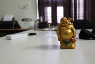 A golden laughing Buddha figurine is placed on a light-colored wooden table. The background includes office items such as a printer, cables, and papers on the table, with dimly lit curtains and a window behind them.