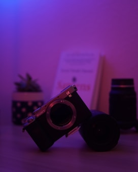 A digital camera body is positioned on its side against a lens, with a softly focused stack of books and a potted plant in the background. Purple and blue lighting creates a dramatic ambiance.