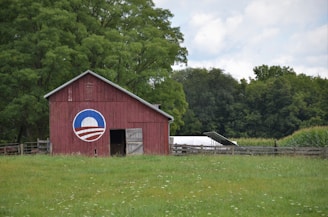A red barn with a white and blue circular symbol painted on it stands in a grassy field. The barn is surrounded by lush green trees and a large section of cornstalks. The sky above is partly cloudy, adding a serene feel to the rural landscape.