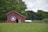 A red barn with a white and blue circular symbol painted on it stands in a grassy field. The barn is surrounded by lush green trees and a large section of cornstalks. The sky above is partly cloudy, adding a serene feel to the rural landscape.