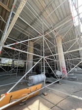 A construction site featuring extensive scaffolding arranged around concrete columns. Construction materials, including large rolls and planks, are scattered on the ground. There are several layers of metal pipes forming a complex network of support structures. The area is partially enclosed, with sunlight streaming through, highlighting the details of the scaffolding. Graffiti is visible on one of the concrete columns.