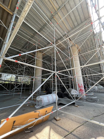A construction site featuring extensive scaffolding arranged around concrete columns. Construction materials, including large rolls and planks, are scattered on the ground. There are several layers of metal pipes forming a complex network of support structures. The area is partially enclosed, with sunlight streaming through, highlighting the details of the scaffolding. Graffiti is visible on one of the concrete columns.