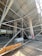 A construction site featuring extensive scaffolding arranged around concrete columns. Construction materials, including large rolls and planks, are scattered on the ground. There are several layers of metal pipes forming a complex network of support structures. The area is partially enclosed, with sunlight streaming through, highlighting the details of the scaffolding. Graffiti is visible on one of the concrete columns.