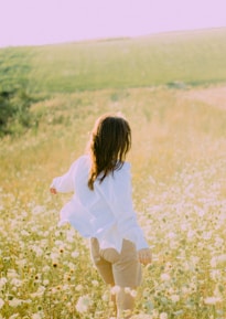 woman in white dress shirt and brown pants walking on yellow flower field during daytime