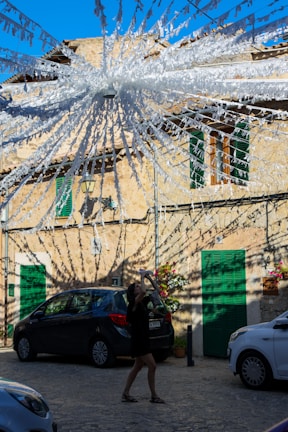 A sunlit cobblestone street with a person taking a photo or video using a smartphone. The street is adorned with white streamers or decorations radiating from a central point above, casting intricate shadows on the buildings and cars. The buildings have stone facades with green shutters, and there are potted plants and flowers adding color to the scene.