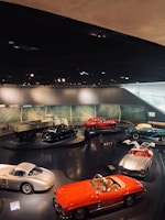 A collection of vintage cars is displayed in an indoor exhibition setting. The cars are parked on a shiny dark floor within a well-lit room, featuring a backdrop of a scenic landscape. The vehicles are various colors such as red, silver, and green. A few people can be seen walking around the display, observing the cars closely.
