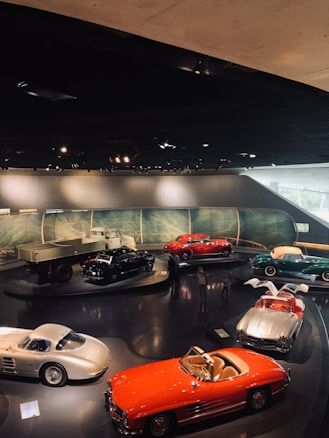 A collection of vintage cars is displayed in an indoor exhibition setting. The cars are parked on a shiny dark floor within a well-lit room, featuring a backdrop of a scenic landscape. The vehicles are various colors such as red, silver, and green. A few people can be seen walking around the display, observing the cars closely.