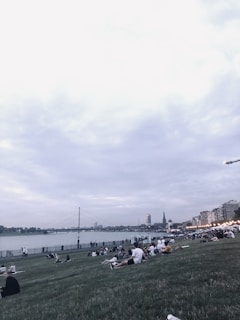 A riverside park scene with people relaxing on a grassy slope. In the background, a large suspension bridge spans across the river, and city buildings can be seen under a cloudy sky. The area is bustling with activity as groups gather on the grass, and lights adorn the riverside path.