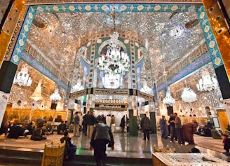 An opulent interior featuring intricate Islamic architecture with detailed mirror work, vibrant blue and gold patterns, and grand chandeliers. People are gathered and sitting on ornate carpets, contributing to a serene and reverent atmosphere.