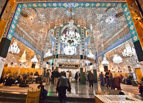 An opulent interior featuring intricate Islamic architecture with detailed mirror work, vibrant blue and gold patterns, and grand chandeliers. People are gathered and sitting on ornate carpets, contributing to a serene and reverent atmosphere.