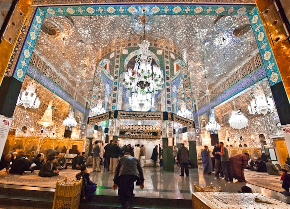An opulent interior featuring intricate Islamic architecture with detailed mirror work, vibrant blue and gold patterns, and grand chandeliers. People are gathered and sitting on ornate carpets, contributing to a serene and reverent atmosphere.