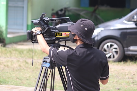 A person wearing a black hat and shirt operates a professional video camera mounted on a tripod outdoors. The surroundings include a parked car and a building with a green wall.