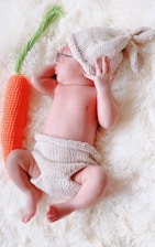 A baby wearing knitted accessories, including a hat and diaper cover, is peacefully lying on a soft, white blanket. Beside the baby is a plush, knitted carrot.