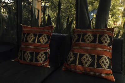 Two patterned cushions with geometric designs rest on a dark fabric sofa. The cushions feature earthy tones, including deep orange and black, complemented by intricate diamond patterns. They are placed against a backdrop of lush greenery and foliage visible through a nearby glass surface.