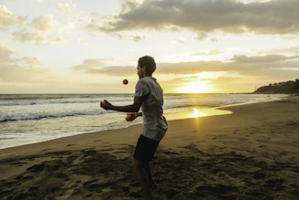 A person is juggling on a beach during sunset, with the ocean waves gently hitting the shore. The sky is filled with a mix of clouds, and the sun casts a warm glow over the scene.