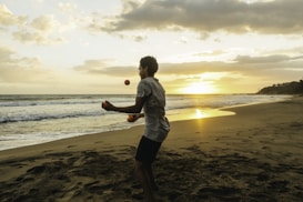 A person is juggling on a beach during sunset, with the ocean waves gently hitting the shore. The sky is filled with a mix of clouds, and the sun casts a warm glow over the scene.