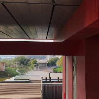 An architectural view capturing a modern building with large red and gray structures framing a scenic outdoor area. The foreground features a series of steps leading to a glass platform, while the background showcases lush greenery and a reflective water body.