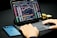 A person is typing on a laptop keyboard while looking at a cryptocurrency trading platform displayed on a tablet screen. Charts and price data are visible. Nearby is a smartphone and a stack of gold coins on the desk.