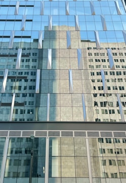 Reflective glass windows of a modern building capturing the distorted reflection of an adjacent structure with rectangular and square patterns. The windows create a geometric visual effect with its repetitive design.