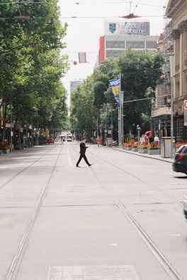 A person in a suit is crossing an empty city street, surrounded by tall trees and buildings. A tramway runs along the street. There are banners on the lampposts and a Bank of Melbourne building in the background.