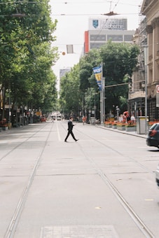 A person in a suit is crossing an empty city street, surrounded by tall trees and buildings. A tramway runs along the street. There are banners on the lampposts and a Bank of Melbourne building in the background.