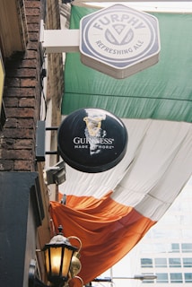 A narrow alley displays multiple signs, including ones for Furphy Refreshing Ale and Guinness. A large fabric with green, white, and orange stripes resembles the Irish flag, draping down the wall. An old-fashioned lantern is affixed to a brick facade, and a portion of a modern building is visible in the background.