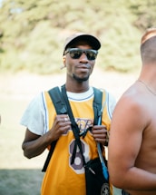 A man wearing sunglasses and a cap is dressed in a yellow sports jersey with the number 8. He has a black backpack and is holding the straps with both hands. Another person, shirtless, is standing beside him with their back partially turned to the camera. The background is outdoors, with blurred greenery.