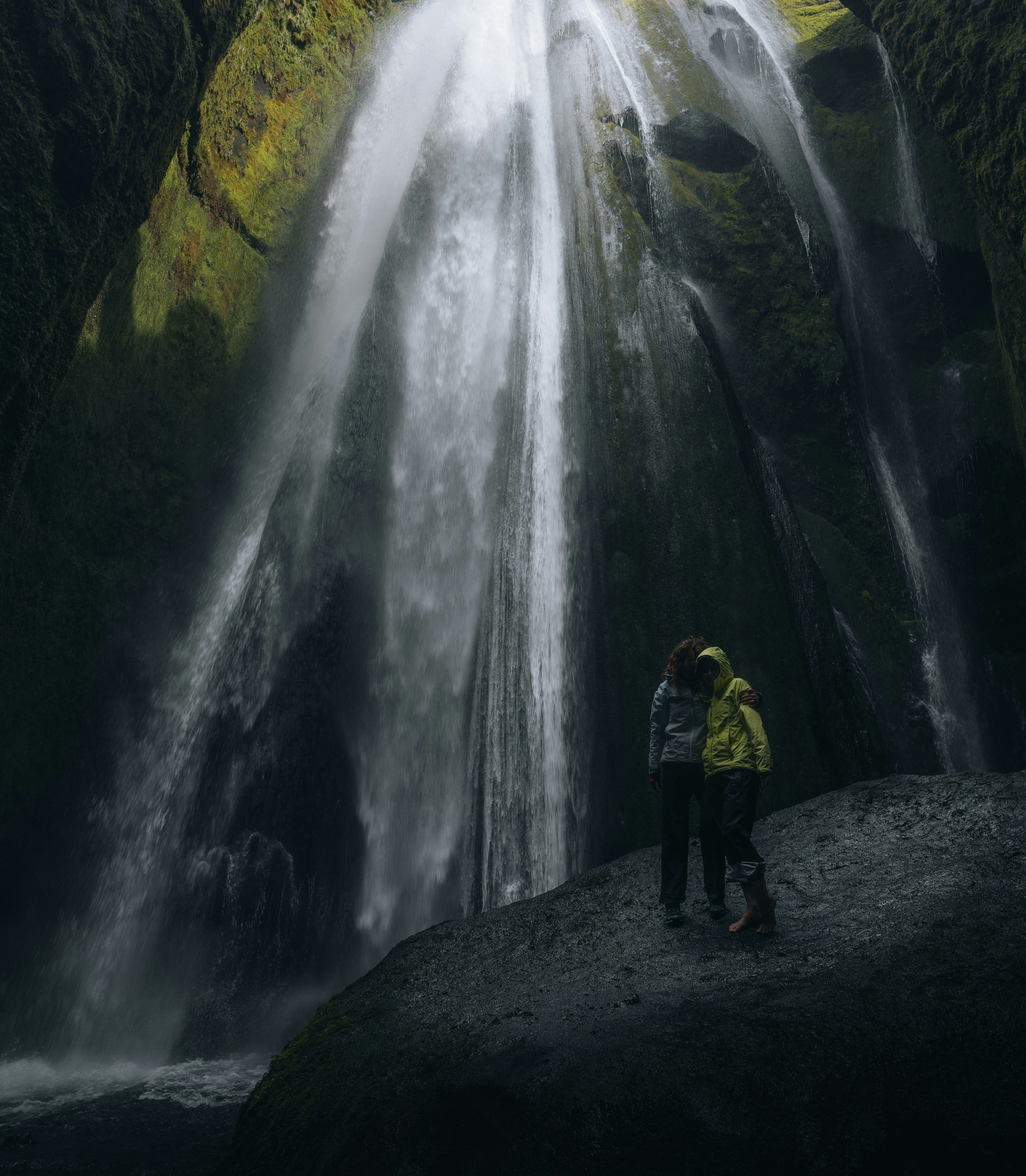 man in yellow jacket and black pants standing on rock near waterfalls during daytime