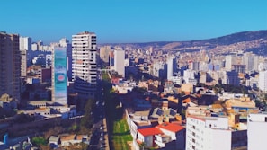 An urban landscape featuring a dense cluster of high-rise buildings. A prominent tower with advertisements stands out among the structures. The scene includes a mixture of residential and commercial buildings, with hills visible in the background under a clear blue sky.