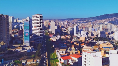 An urban landscape featuring a dense cluster of high-rise buildings. A prominent tower with advertisements stands out among the structures. The scene includes a mixture of residential and commercial buildings, with hills visible in the background under a clear blue sky.