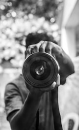A person is holding a camera lens close to the viewer, focusing on the circular shape and reflective surface of the lens. The background is blurred, creating a depth-of-field effect, with light and shadow creating a patterned texture.