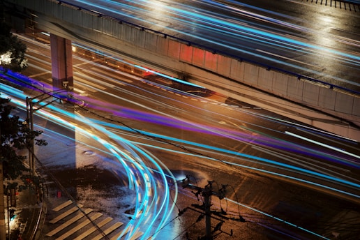 Long-exposure photograph capturing an urban street scene at night with streaks of light from moving vehicles. The image prominently features a wet road surface reflecting light, an overpass, and various street elements such as a zebra crossing and traffic light poles.
