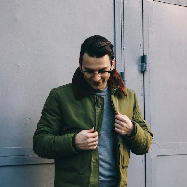 A man in a green jacket standing near a white wall