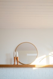 A minimalistic interior scene featuring a round mirror with a thin wooden frame, placed on a wooden shelf against a light wall. Two wooden candlestick holders with white candles on top are positioned to the left of the mirror. Sunlight casts soft shadows on the wall, and a green plant leaf is visible on the right.