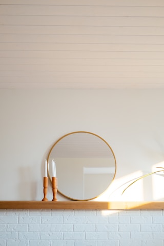 A minimalistic interior scene featuring a round mirror with a thin wooden frame, placed on a wooden shelf against a light wall. Two wooden candlestick holders with white candles on top are positioned to the left of the mirror. Sunlight casts soft shadows on the wall, and a green plant leaf is visible on the right.