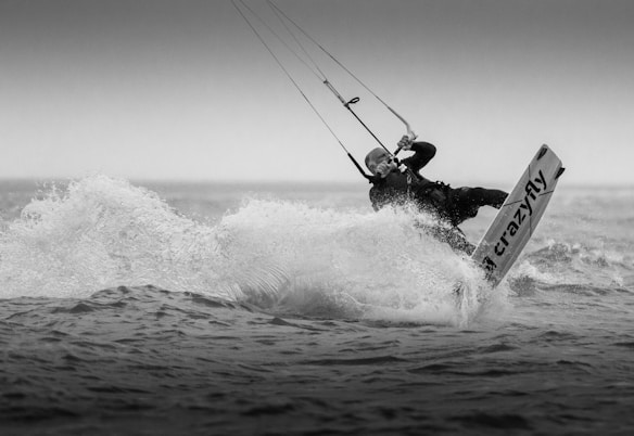 A person is engaged in kite surfing, captured mid-action as they ride the waves. The image is in black and white, focusing on the dynamic movement and the splashing water around the kite surfer.