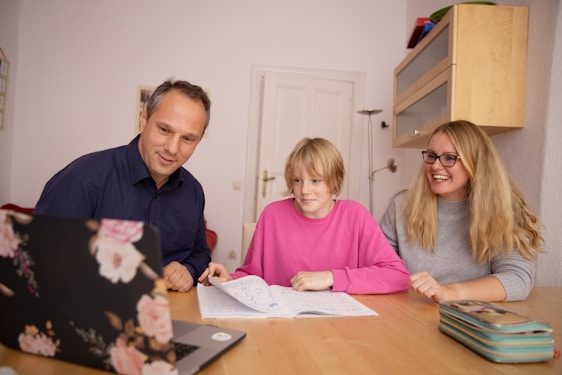 A family scene with three people gathered around a wooden table, two adults and a child. They are engaged with a laptop that features a floral design, and an open notebook is on the table. The atmosphere appears to be positive and collaborative.