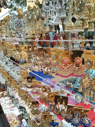 A vibrant display of various traditional earrings with intricate designs in gold, silver, and colorful beads and stones. The earrings are neatly organized on a market stall, with labels indicating they are fashion accessories. The background hints at an outdoor market setting with blurred figures and textiles.