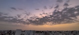 A skyline featuring a cityscape with numerous buildings under a dramatic, cloudy sky. The clouds are scattered across the horizon as the sky transitions from a pale blue to a warm yellow, suggesting either sunrise or sunset.
