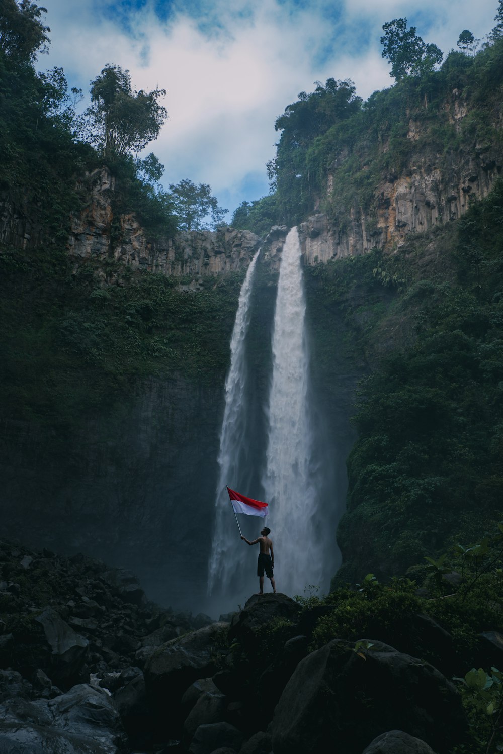 person in red jacket standing on rock near waterfalls during daytime