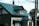 A residential house with a distinctive blue-green metal roof. The structure includes multiple stories and windows, with visible chimneys and a flag attached to the side. Overhead cables and poles are present in the foreground, adding a suburban or urban feel to the scene. The lighting suggests late afternoon with shadows cast on the walls.