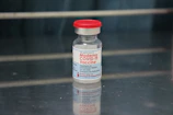 A vial labeled 'Moderna COVID-19 Vaccine' with a red cap. It is placed on a reflective metallic surface, with an out-of-focus background. The vial contains a clear liquid and detailed instructions are visible on the label.