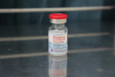 A vial labeled 'Moderna COVID-19 Vaccine' with a red cap. It is placed on a reflective metallic surface, with an out-of-focus background. The vial contains a clear liquid and detailed instructions are visible on the label.