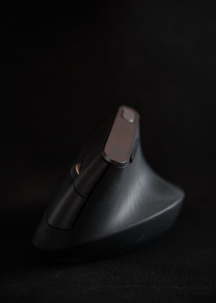 A black ergonomic computer mouse with a vertical design is set against a dark background. The mouse features a sleek, modern shape with multiple buttons visible. It appears angled slightly away, with a matte finish and a smooth texture.